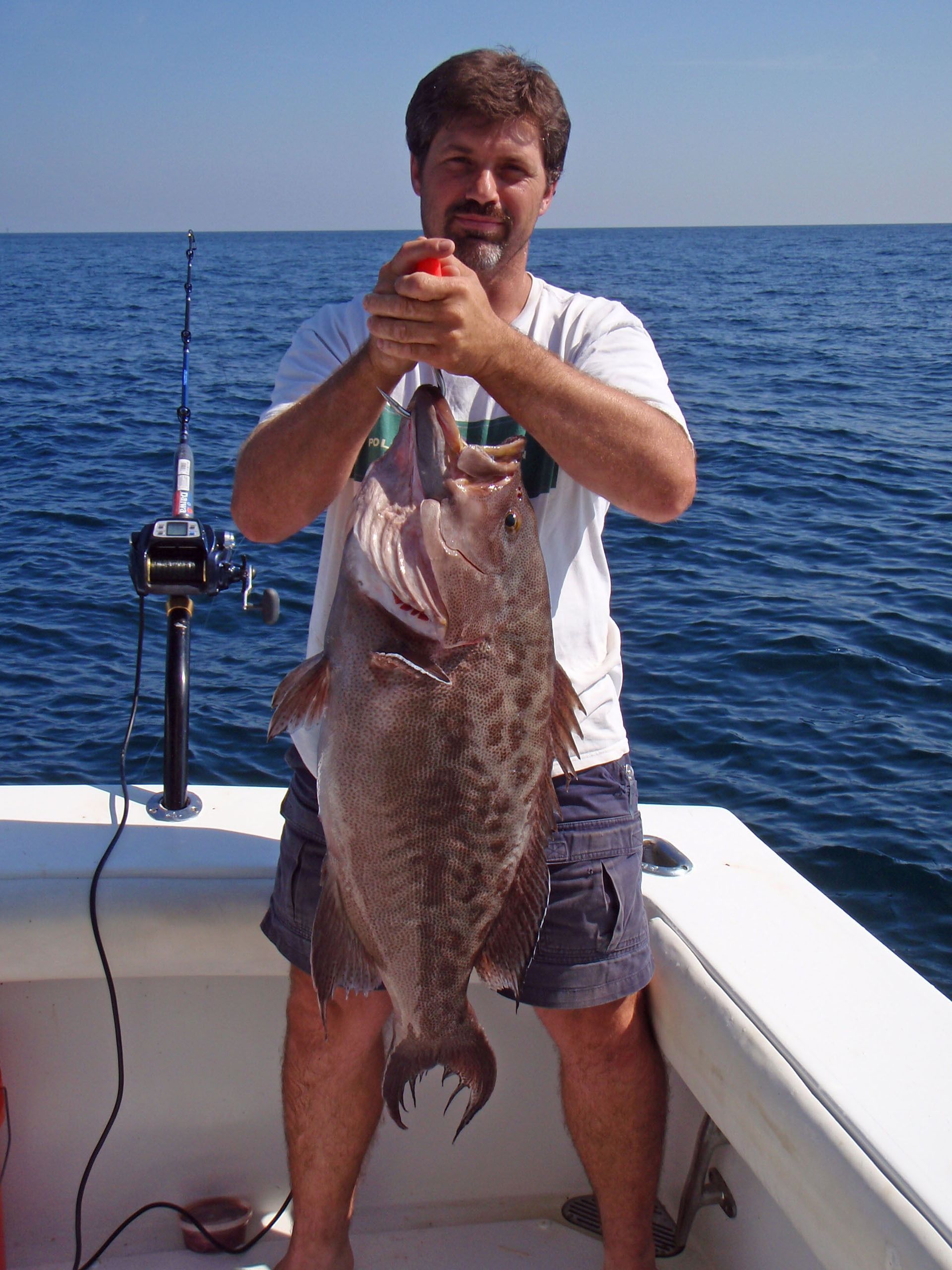 A person standing on a boat holds a large, brown speckled fish with both hands against a backdrop of the open blue sea.