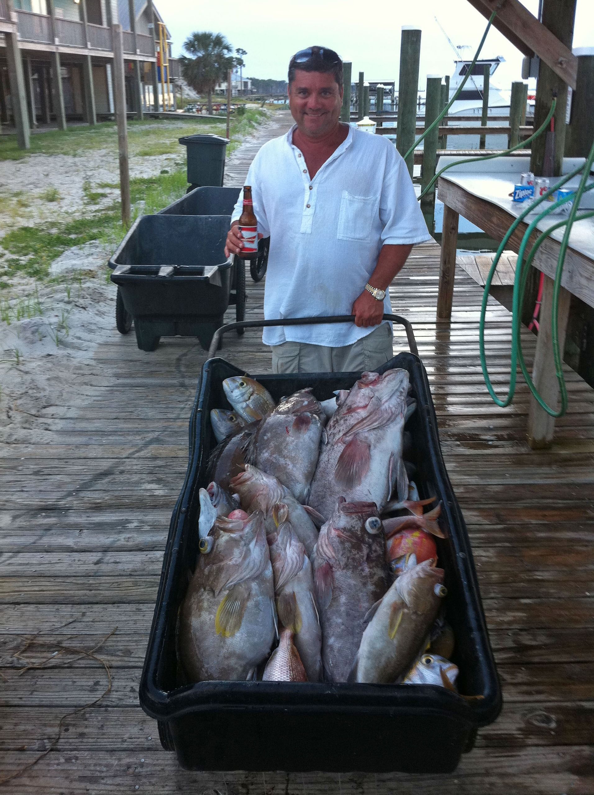A person stands on a wooden dock holding a drink, with a large, full crate of fresh-caught fish in front of them.