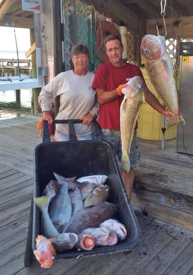 Two people on a wooden dock stand behind a wheelbarrow full of fish; one holds a large fish suspended from a hook.