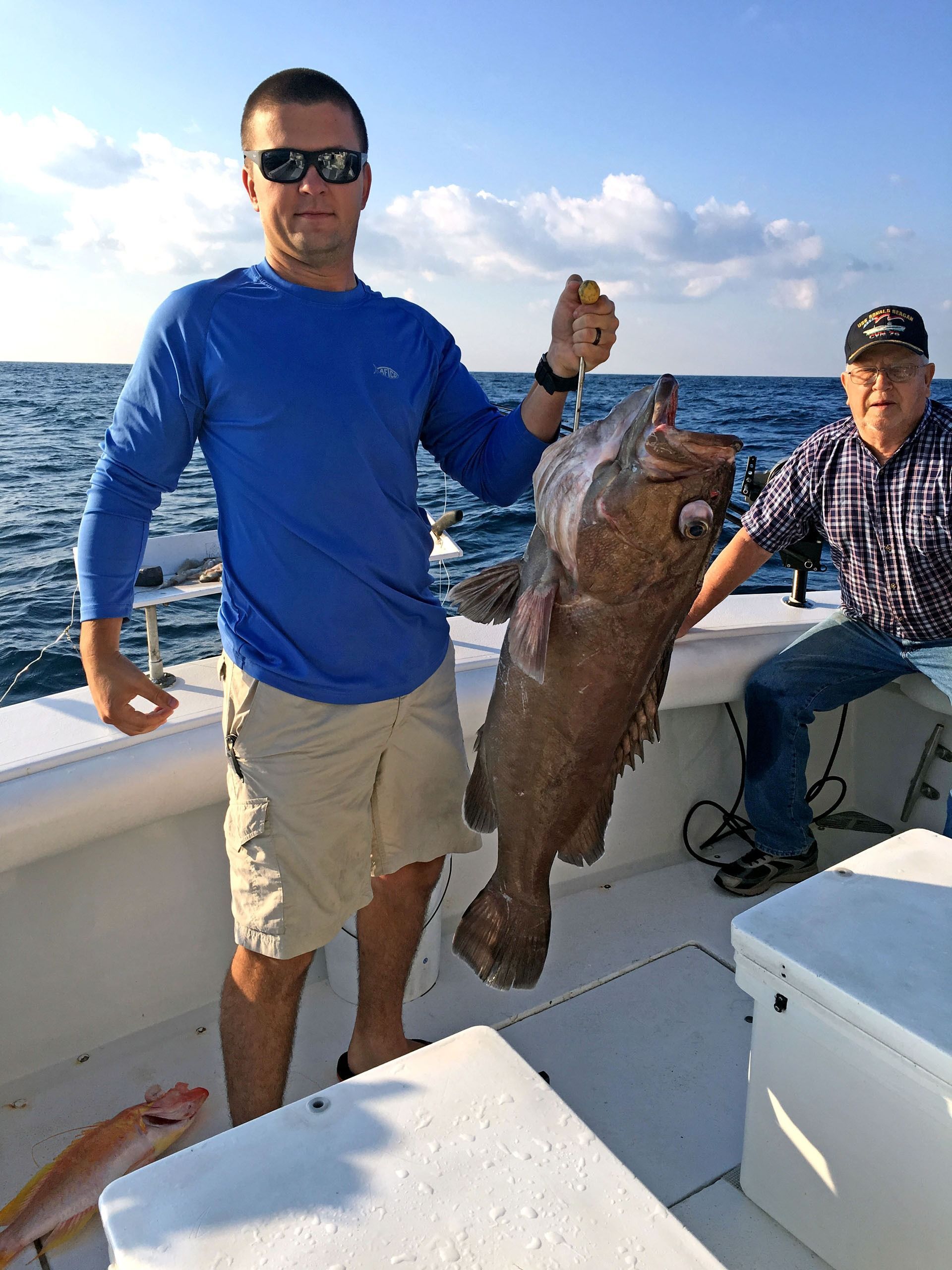 A person in a blue long-sleeve shirt holding a large fish on a boat, with another person sitting nearby.