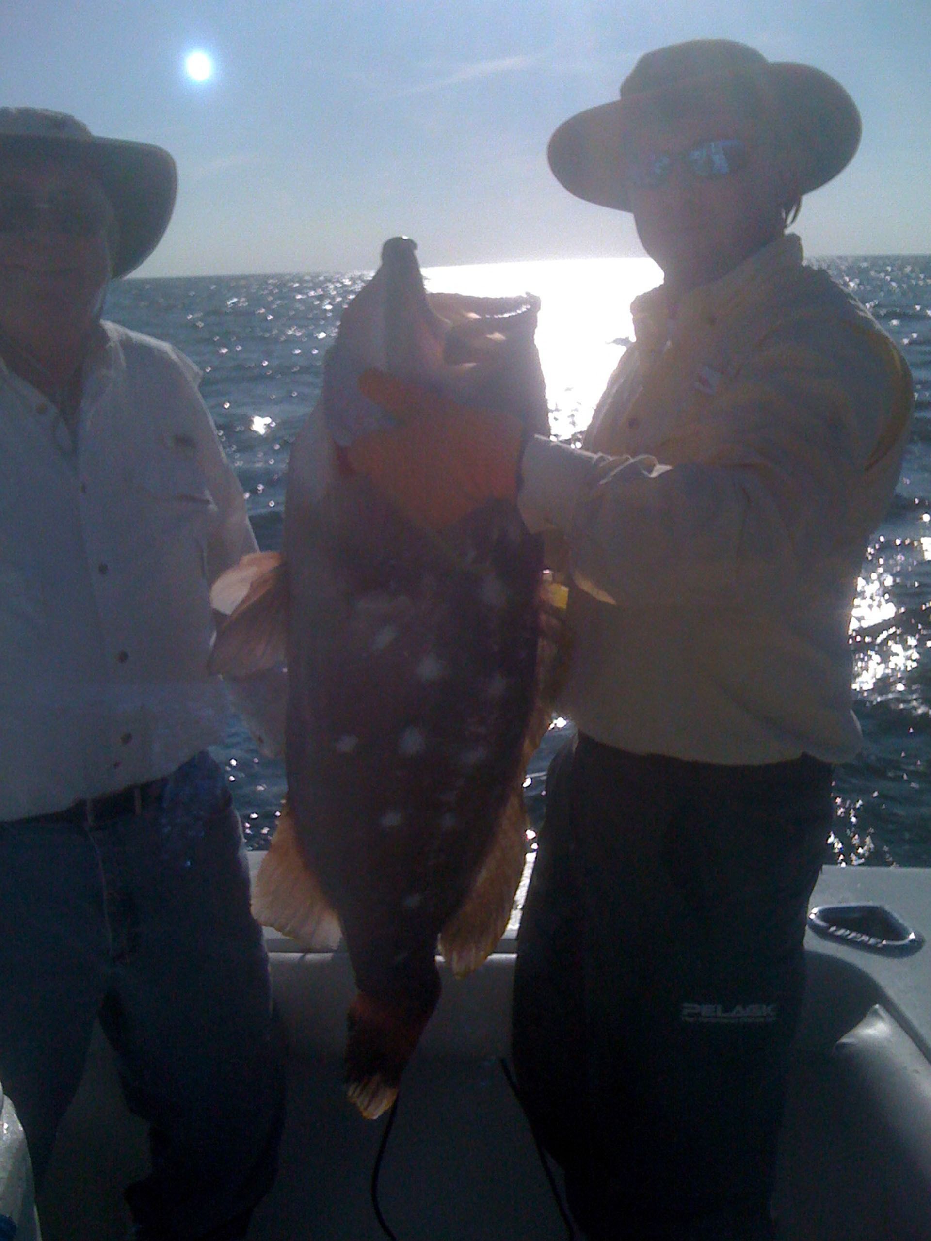 Two people in hats on a boat holding a large, speckled fish up for a photo against a bright, sunny ocean background.