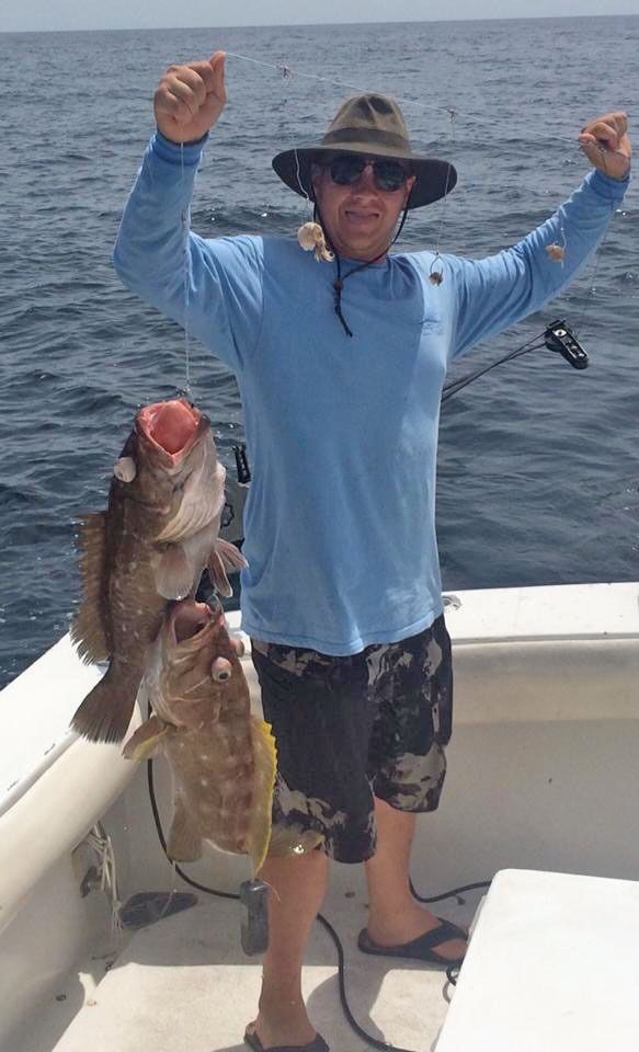 A person on a boat wearing a hat and sunglasses, smiling and holding up two caught fish.
