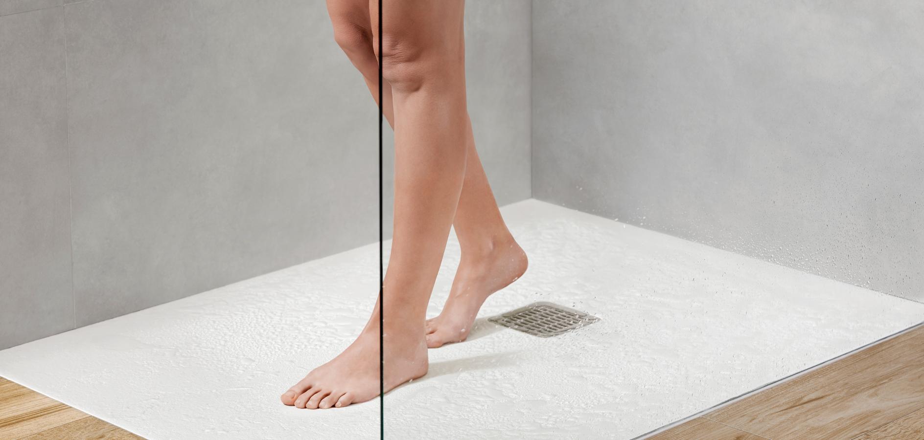 Person's legs and feet in a shower, stepping onto a textured white surface, neutral gray walls.