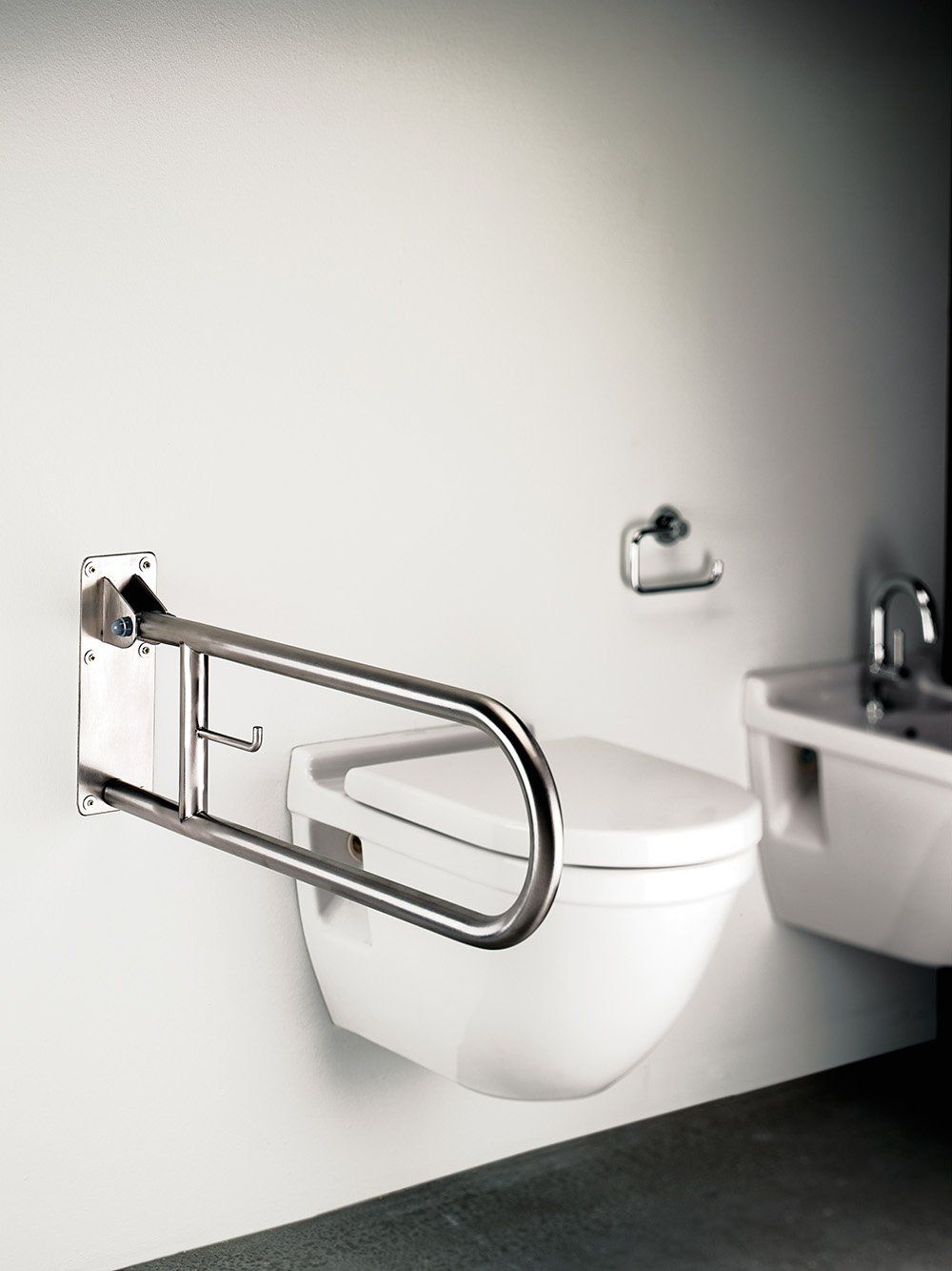 Wall-mounted toilet with folding grab bar in accessible bathroom. White toilet, silver bar, white wall.