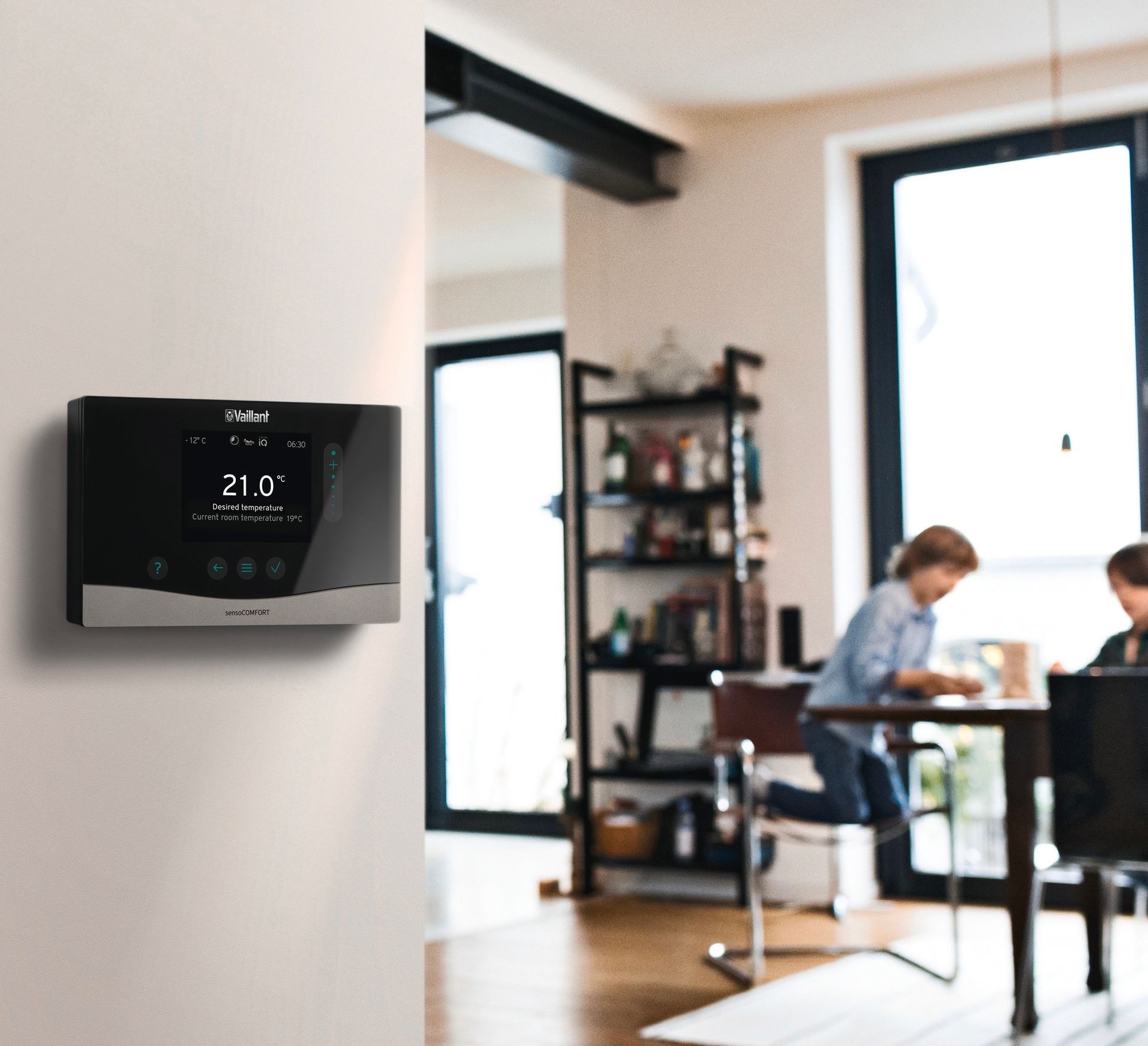 Thermostat showing 21°C on a white wall. Kids play in the background near a bookcase and table.