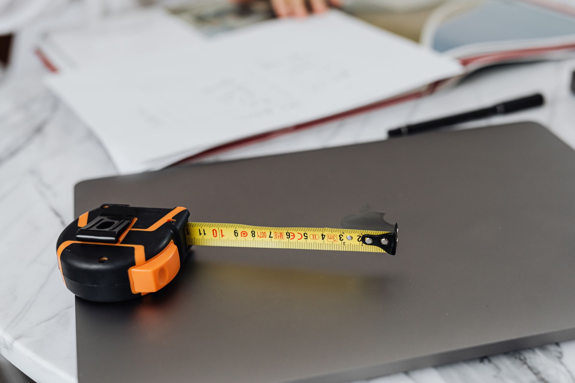 Tape measure extended on closed laptop. Notebook and pen in the background.