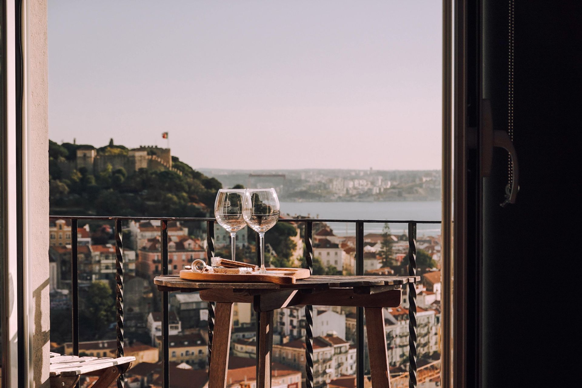 Two wine glasses are on a table on a balcony overlooking a city.