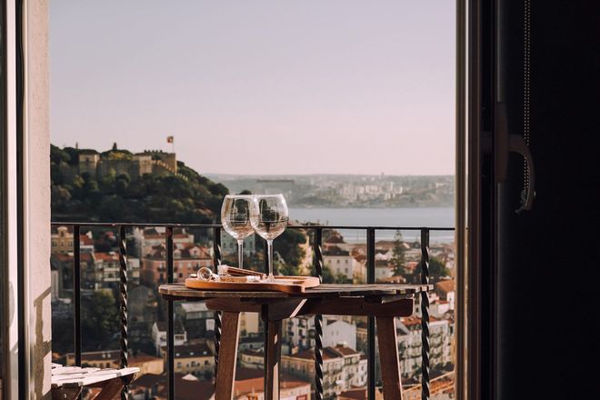 Two wine glasses are on a table on a balcony overlooking a city.