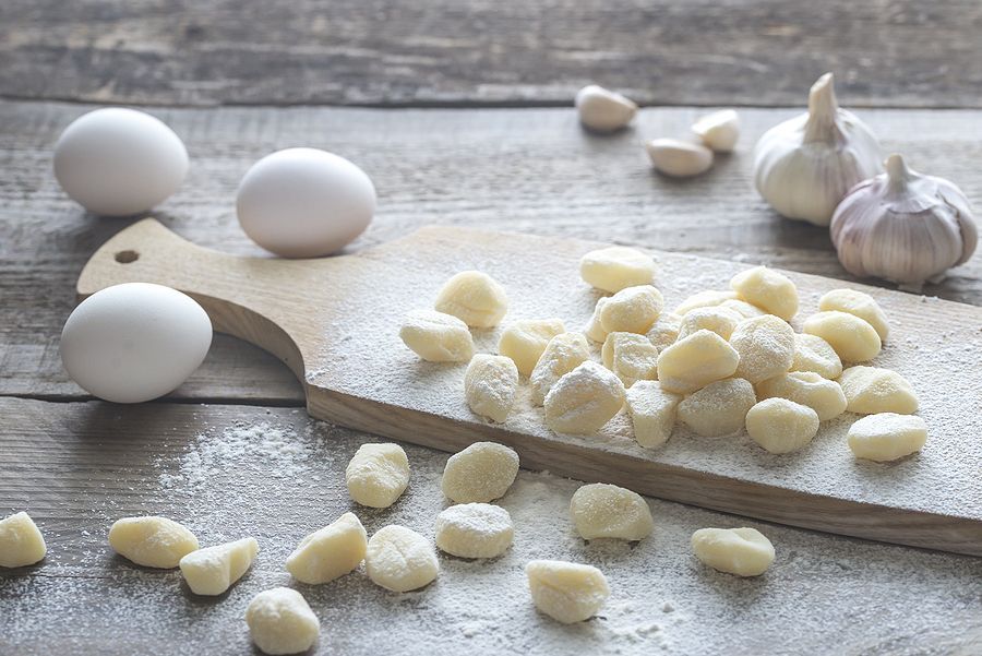 Gnocchi on a cutting board next to eggs and garlic on a wooden table.