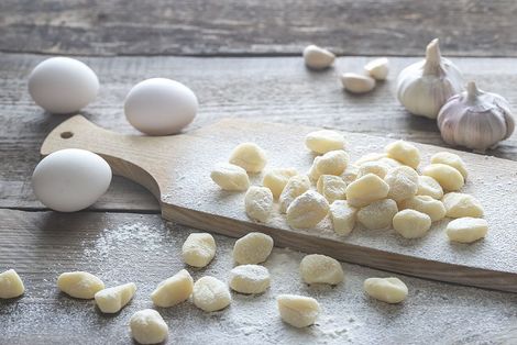 Gnocchi on a cutting board next to eggs and garlic on a wooden table.