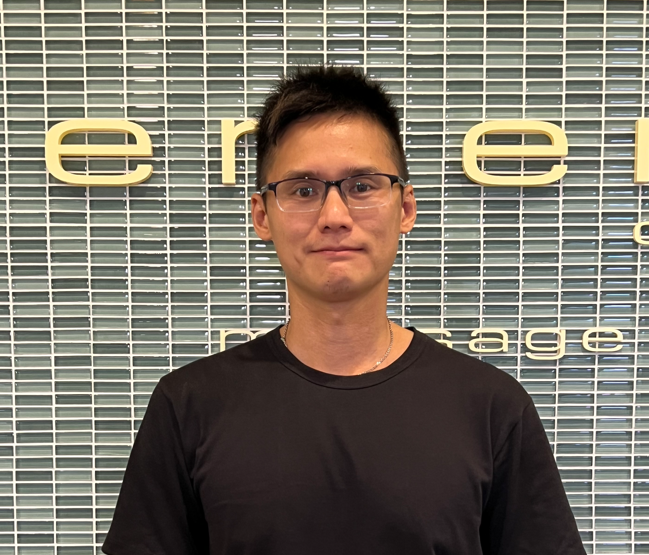 A man wearing glasses and a black shirt is standing in front of a tiled wall.