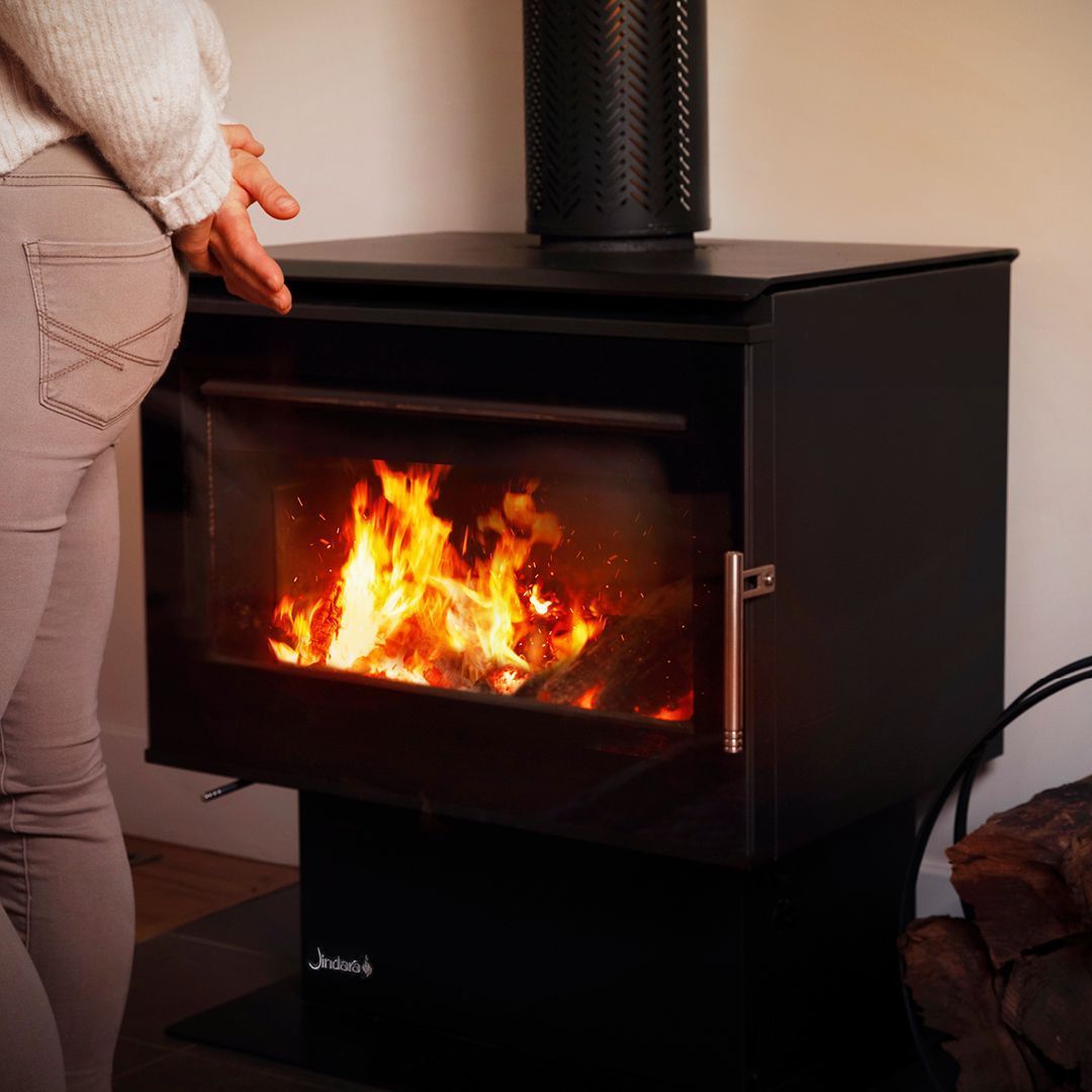 A woman is standing in front of a wood stove with a fire coming out of it.