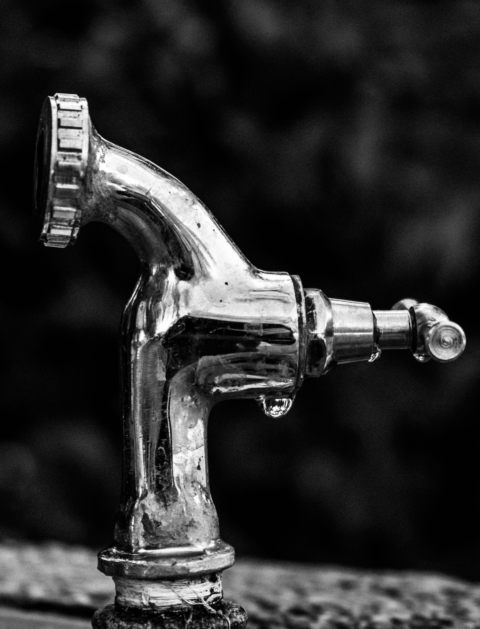 Black and white photo of a shiny silver water faucet, with a water droplet falling from the spout.
