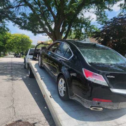 Black Acura sedan on a tow truck, parked on a street under a tree on a sunny day.