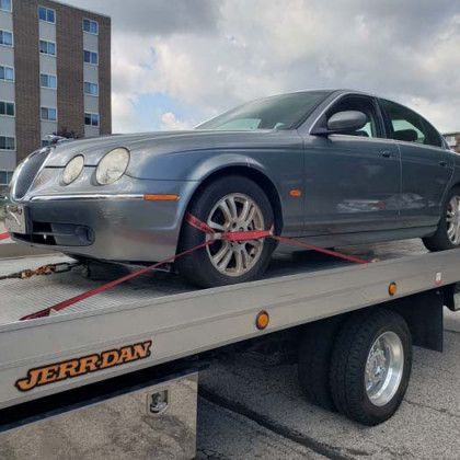 A gray Jaguar S-Type sedan being towed on a flatbed tow truck on a cloudy day.