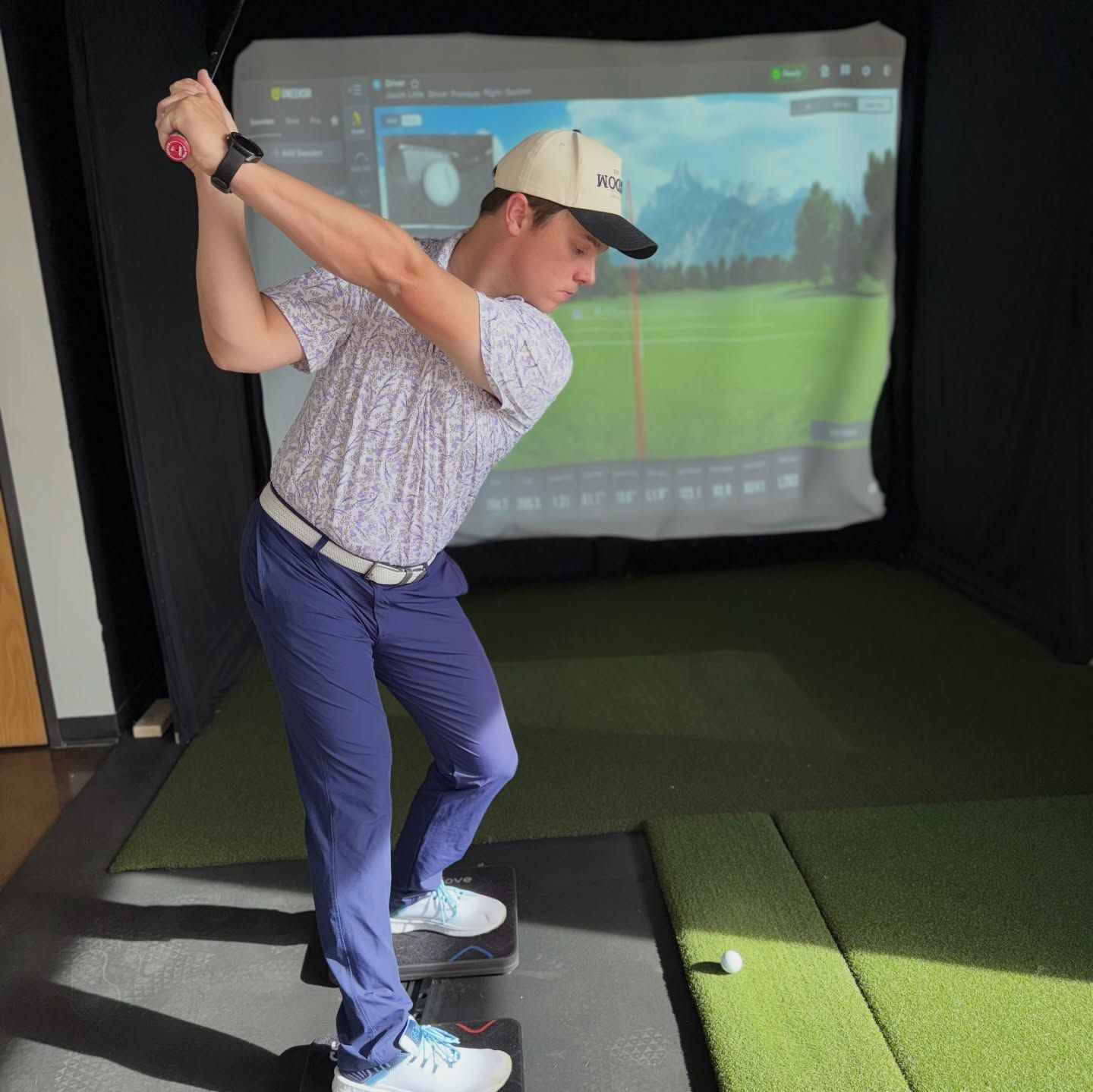 Golfer mid-swing in an indoor golf simulator, wearing a cap and blue pants.