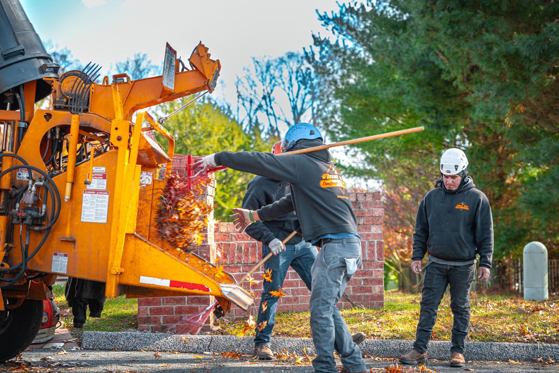 Three workers feed branches into an orange wood chipper on a sunny day.