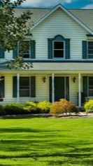 Two-story house with green shutters, a porch, and a well-manicured lawn under a sunny sky.