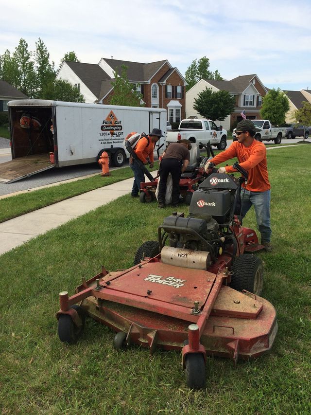 Three landscapers tending to equipment on a lawn near a trailer and houses.