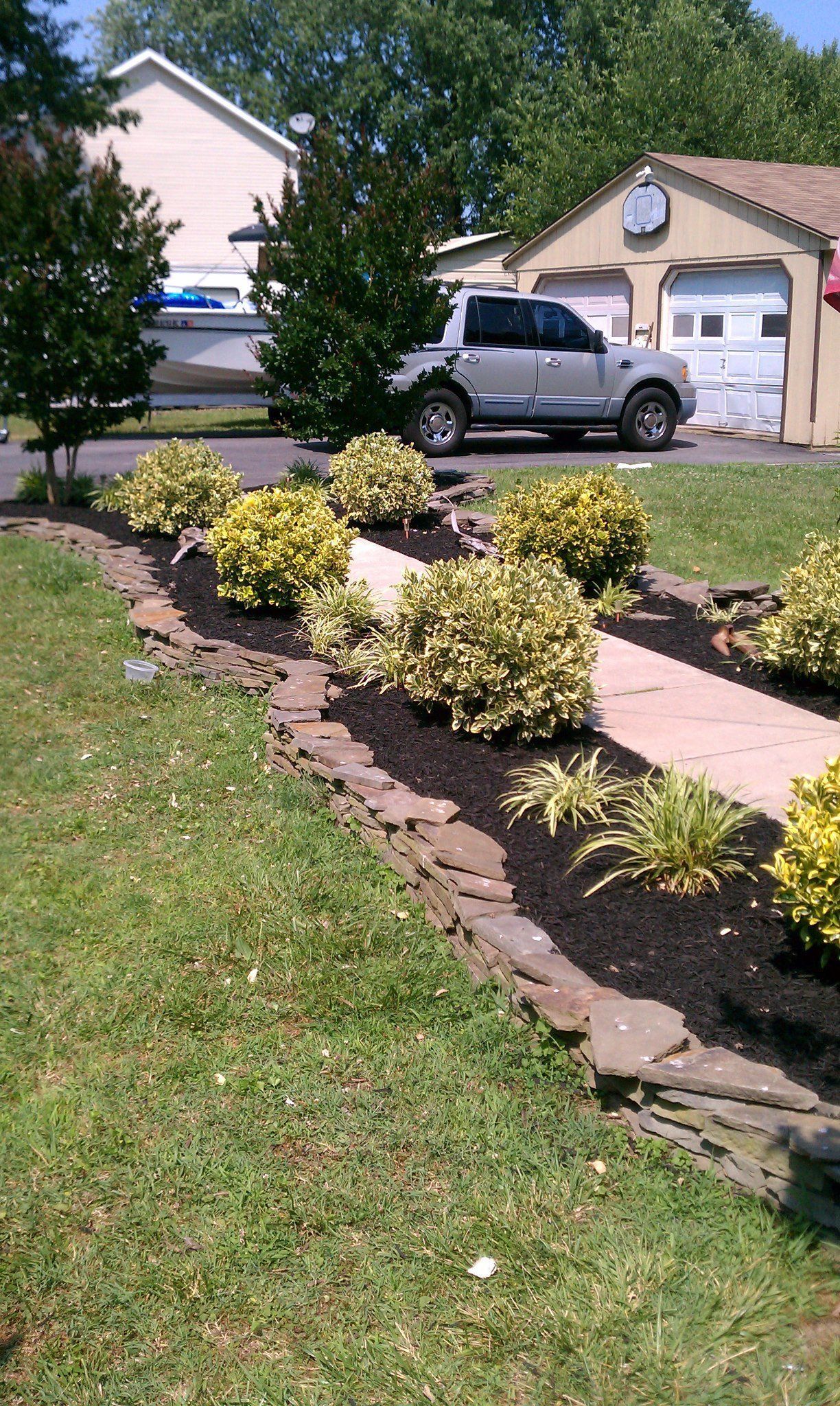 Landscaped yard with stone edging, mulch, yellow bushes, and a pathway to a gray truck and garage.