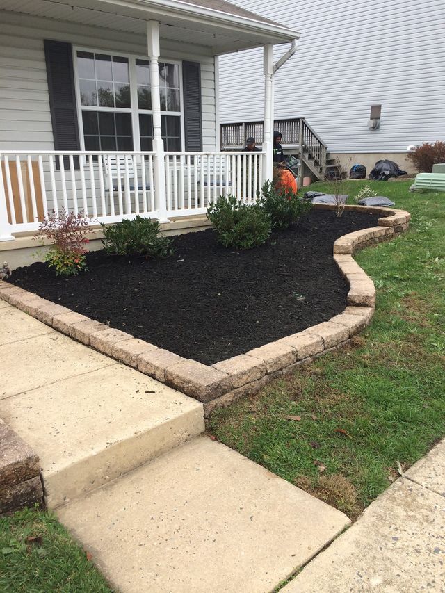 Black mulch in a landscaped bed edged with stone blocks; white porch railing; concrete walkway.