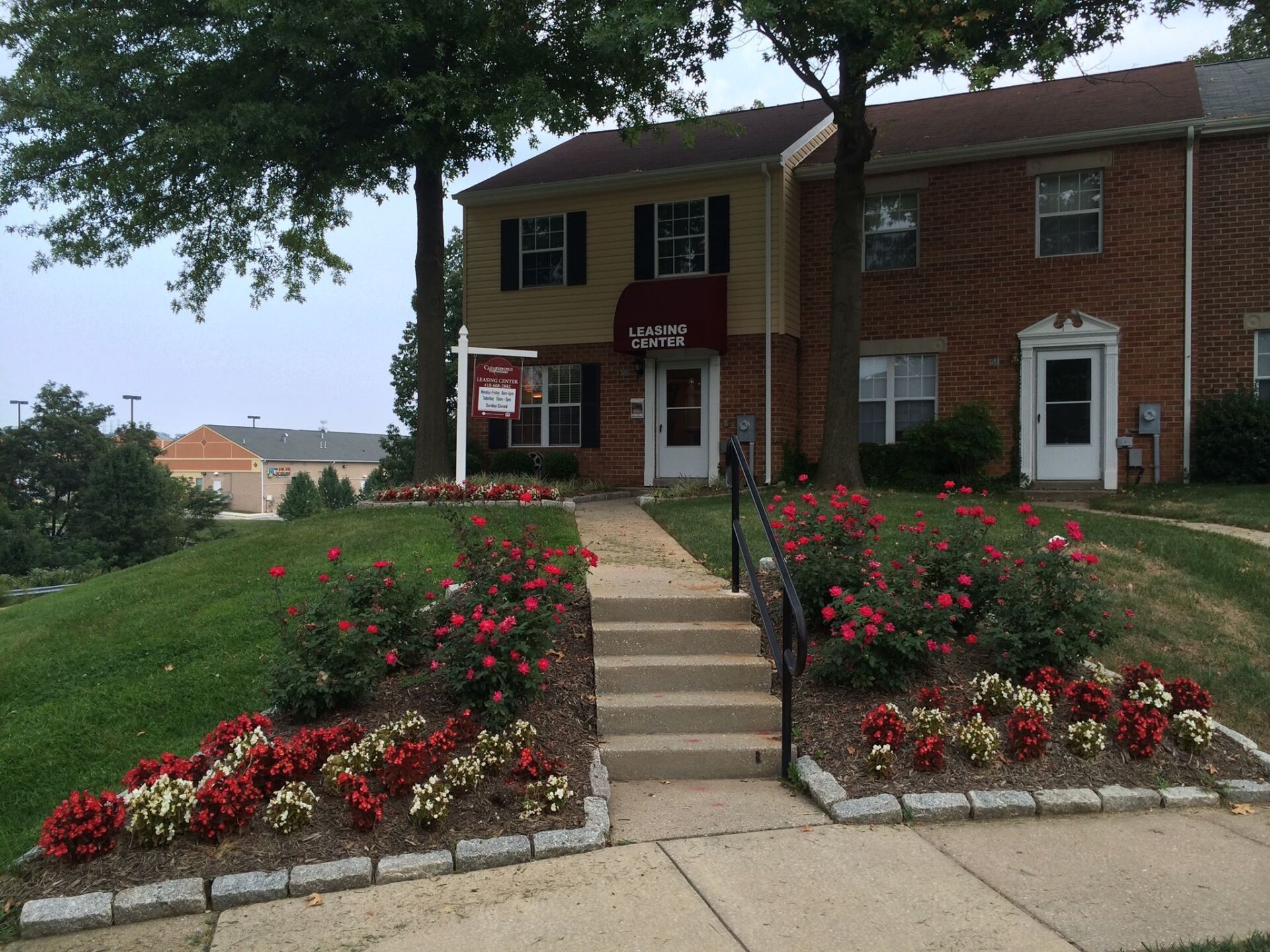 Townhouse exterior with steps, landscaping, and a for-sale sign. Brick and tan siding, red and white flowers.