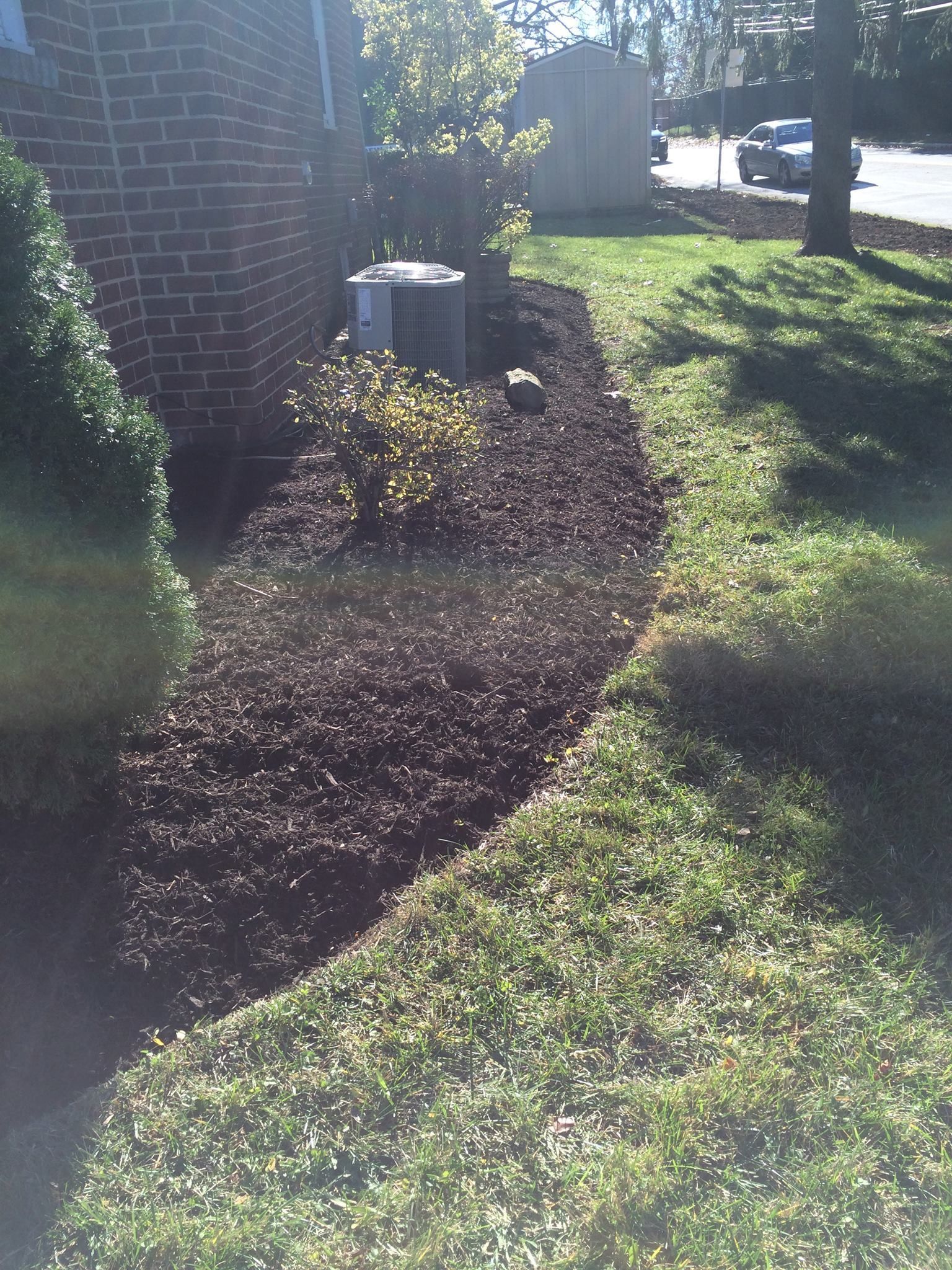 Landscaped yard bed with dark mulch and plants, next to green lawn, brick house, and AC unit.