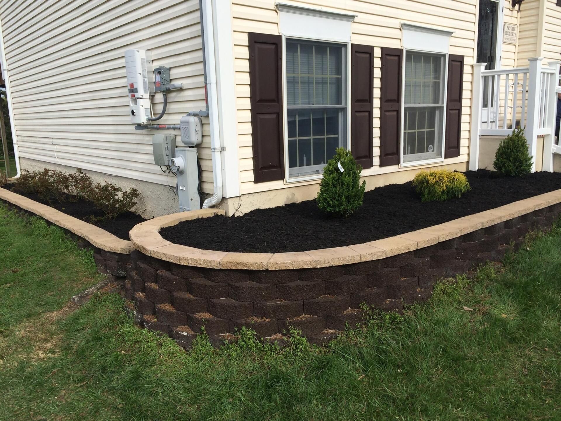 Brown brick retaining wall with mulch and shrubs in front of a house.