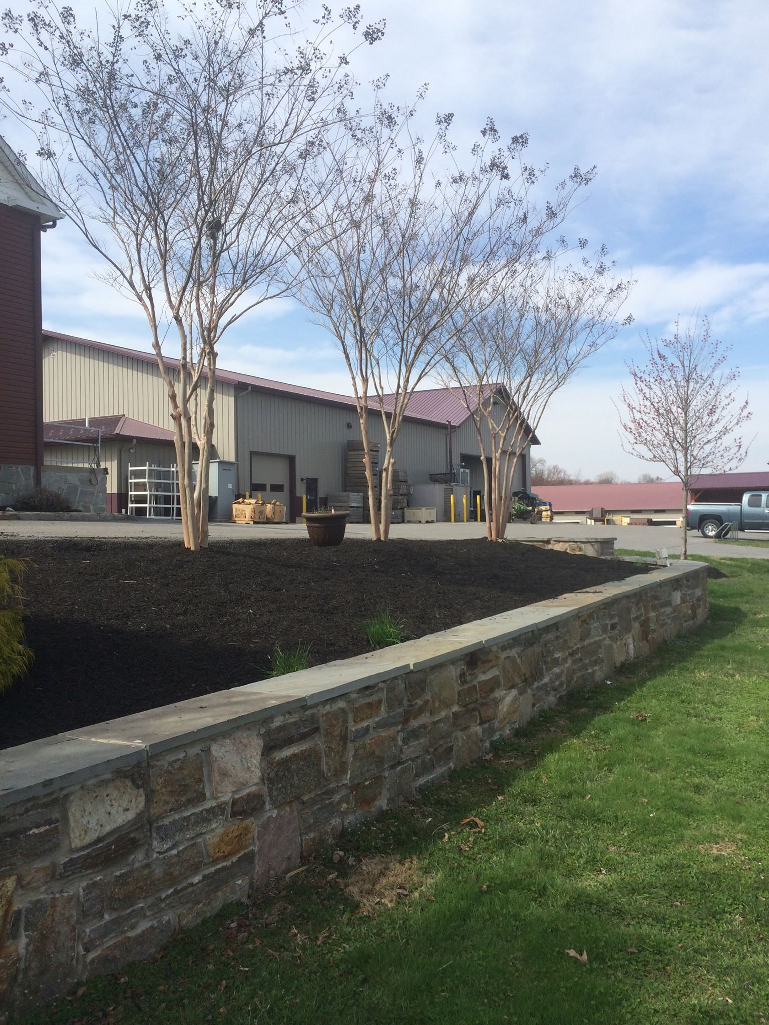 Stone wall in front of dark mulch and trees. Building with red roof and silo in the background. Sunny day.