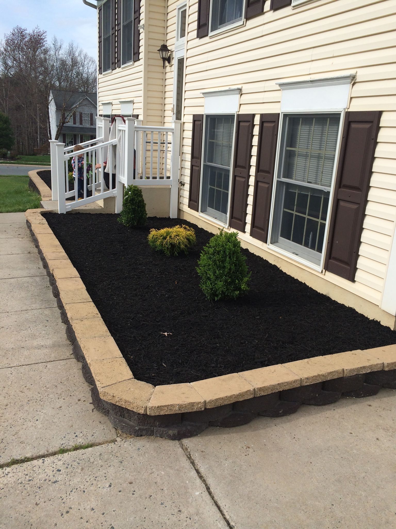 Black mulch bed with beige retaining wall edging in front of a beige house with brown shutters.