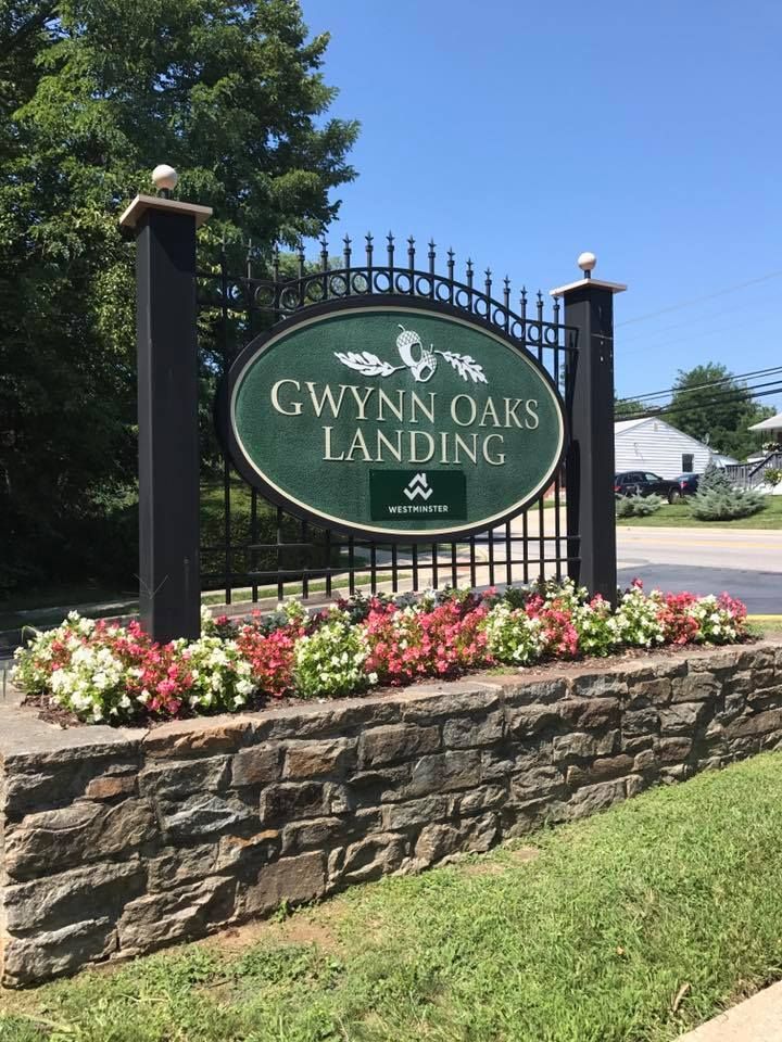 Gwyn Oaks Landing sign with green and white flowers, black metal, and a stone wall.