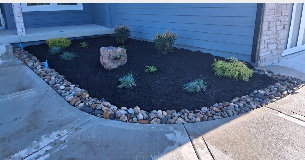 Landscaped corner bed with dark mulch, small shrubs, rocks, and a curved stone border beside a house walkway