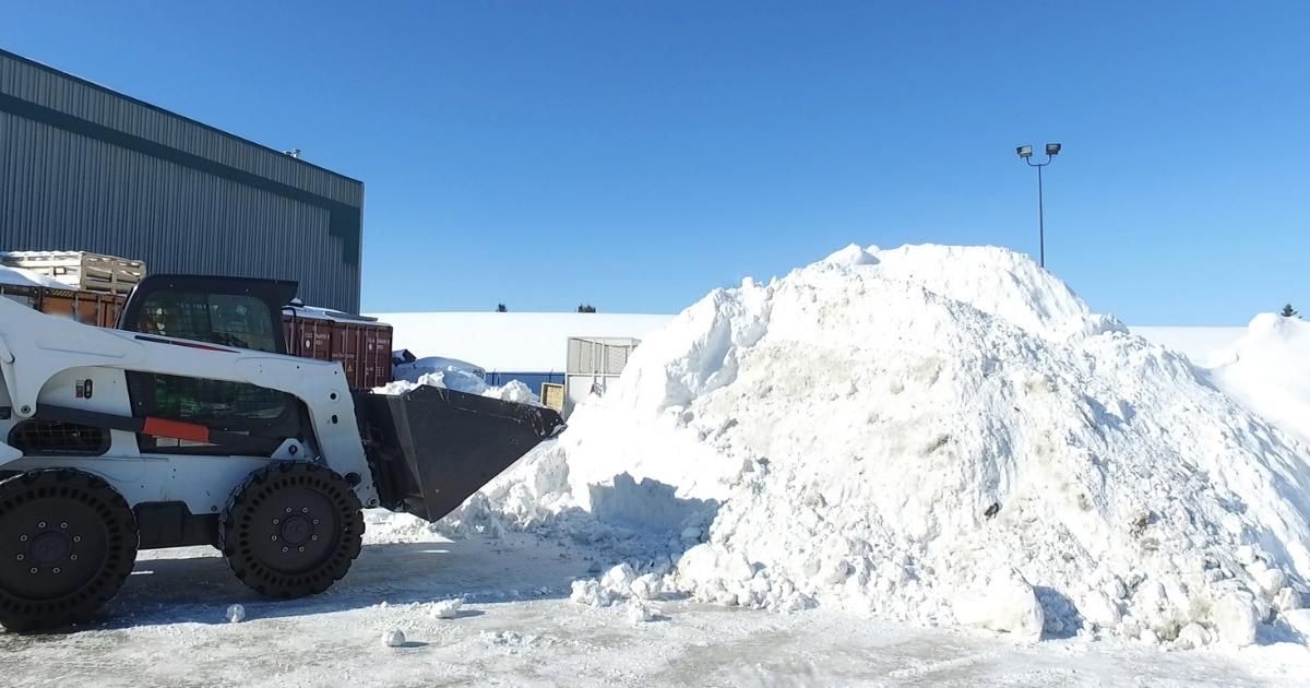 Skid-steer loader scoops snow into a large pile beside a building under a clear, blue sky.
