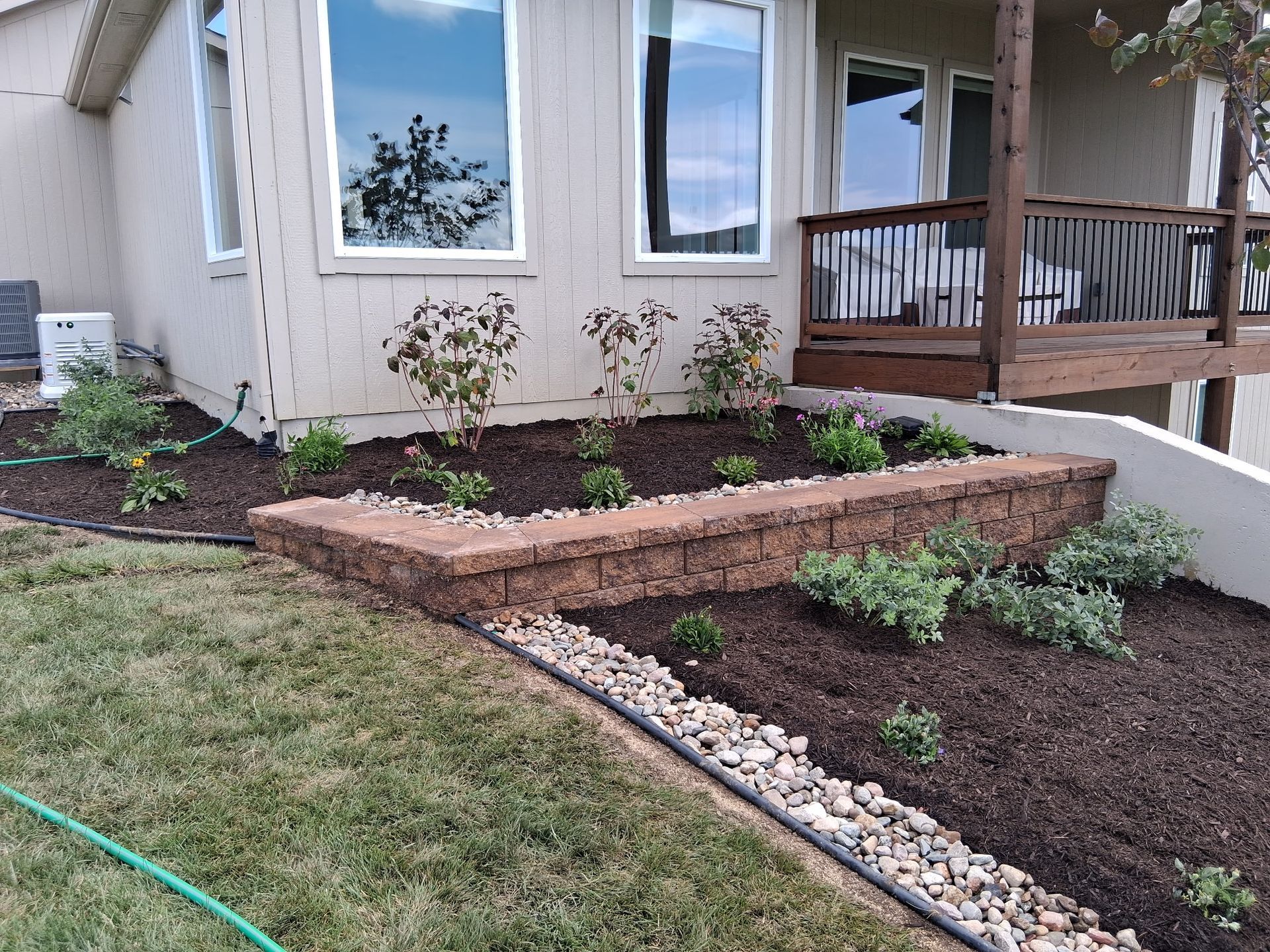 A retaining wall with plants in front of a house, surrounded by brown mulch and green grass.