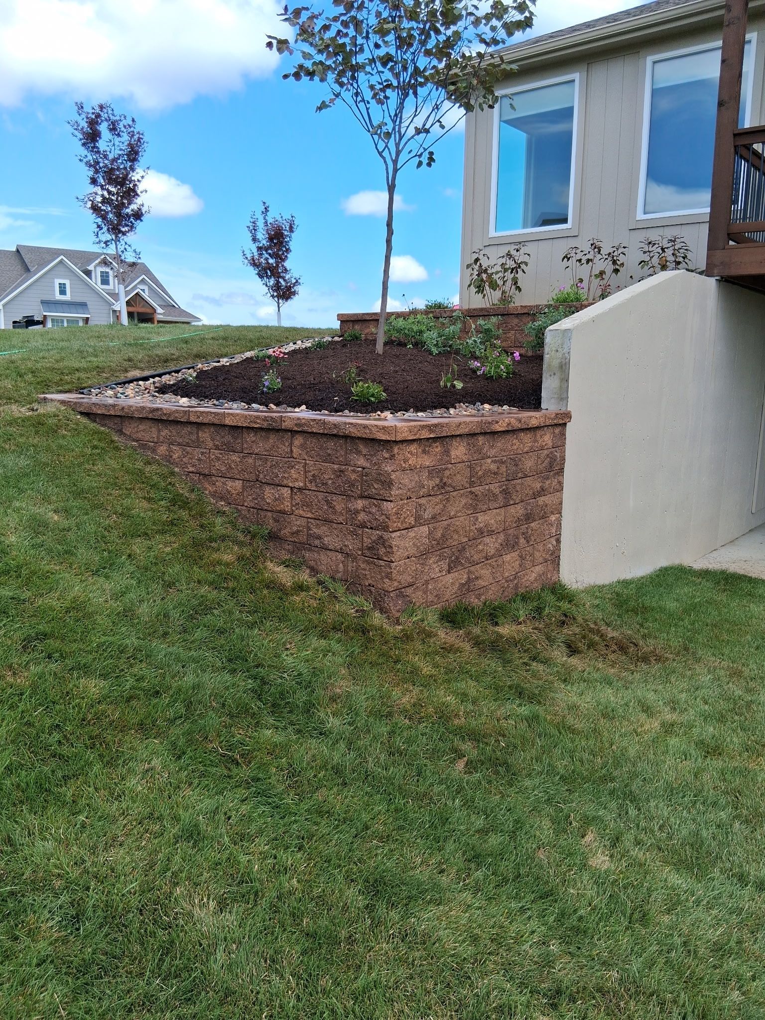 A retaining wall made of brown bricks supports a planted garden bed near a house, on a grassy hillside.