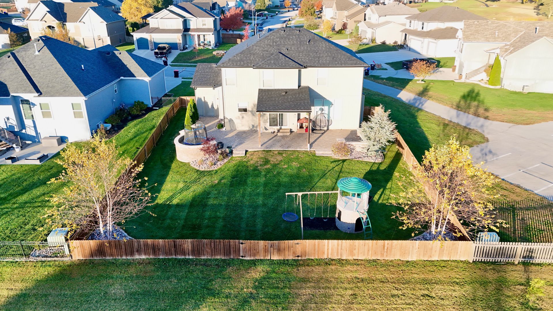Aerial view of a suburban house with a backyard. Green lawn, wooden fence, play structure, patio, and trees.