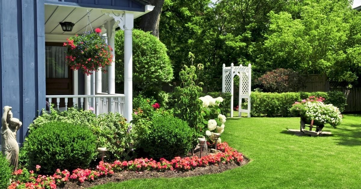 Blue house with a lush green lawn and garden. Red and white flowers, white trellis, and statue.