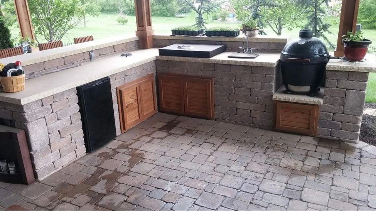 Outdoor kitchen with stone counters, built-in grill, sink, refrigerator, and wooden cabinets, on a stone patio.