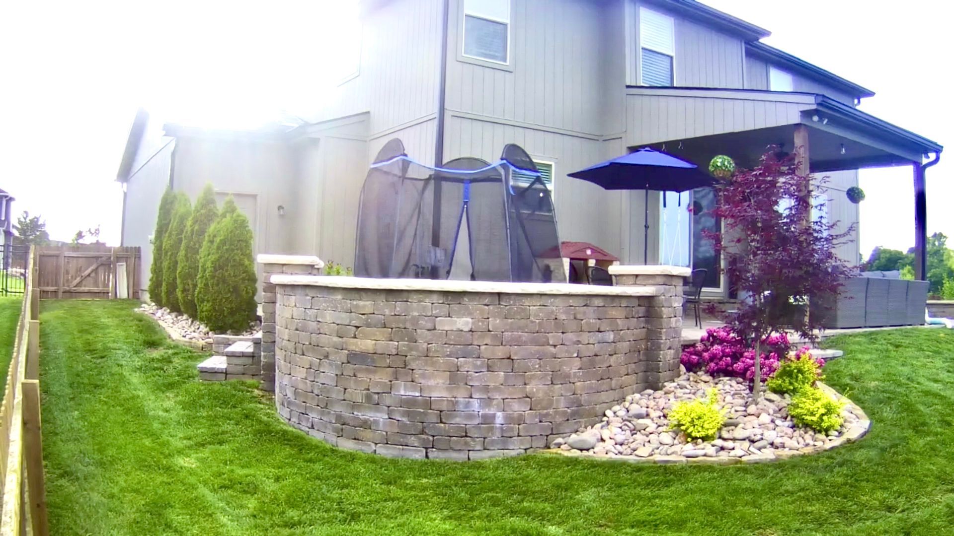 Backyard patio with stone wall, trampoline, and landscaping in front of a house.