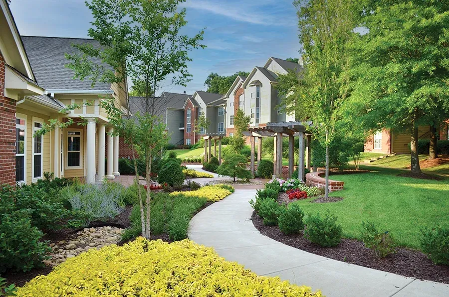 A winding concrete path leads through landscaped green lawns and gardens toward apartment buildings on a sunny day.