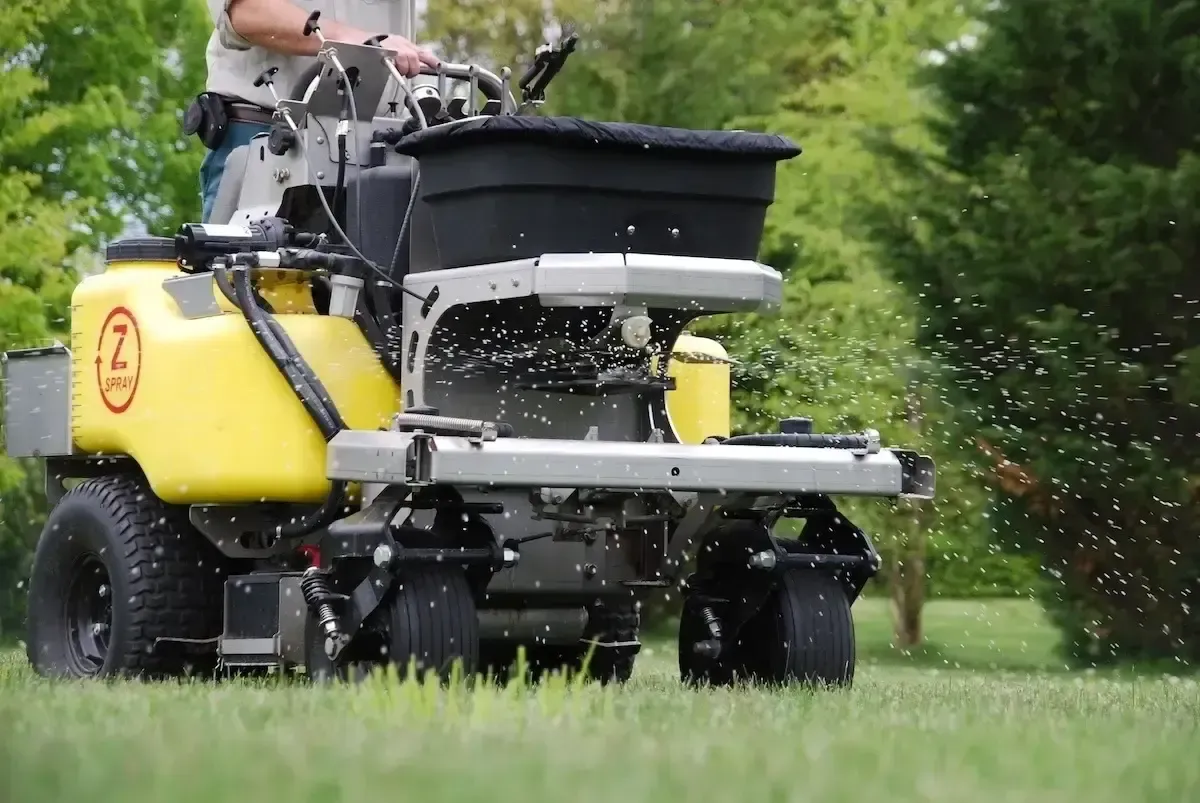 A person operates a yellow Z-Spray spreader-sprayer machine as it disperses fertilizer granules across a grassy lawn.