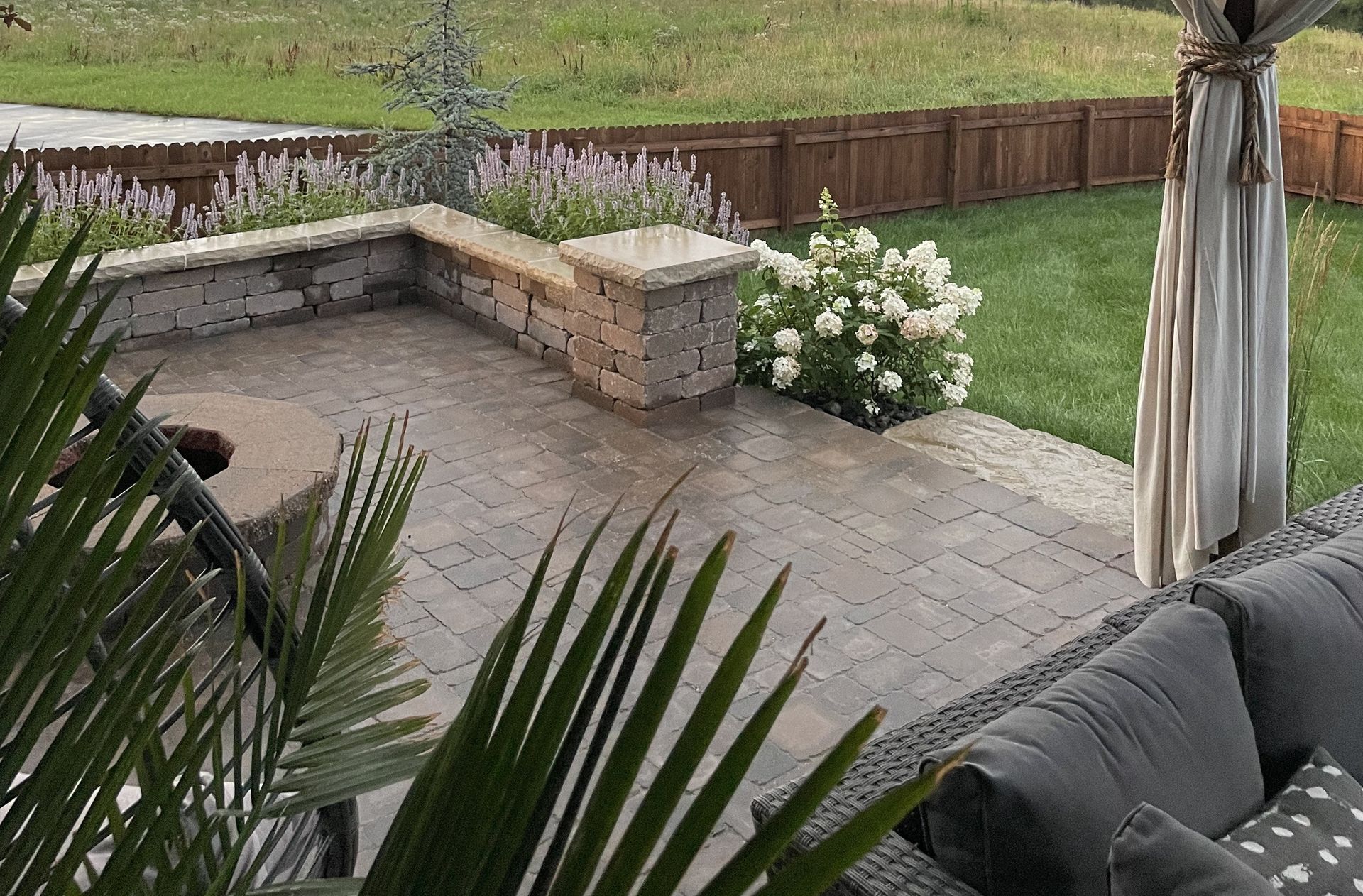 A paved patio with a stone fire pit, seating area, and flower garden, overlooking a grassy yard with a wooden fence.