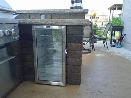 Outdoor kitchen with brick enclosure, grill, and stainless steel refrigerator on a patio.