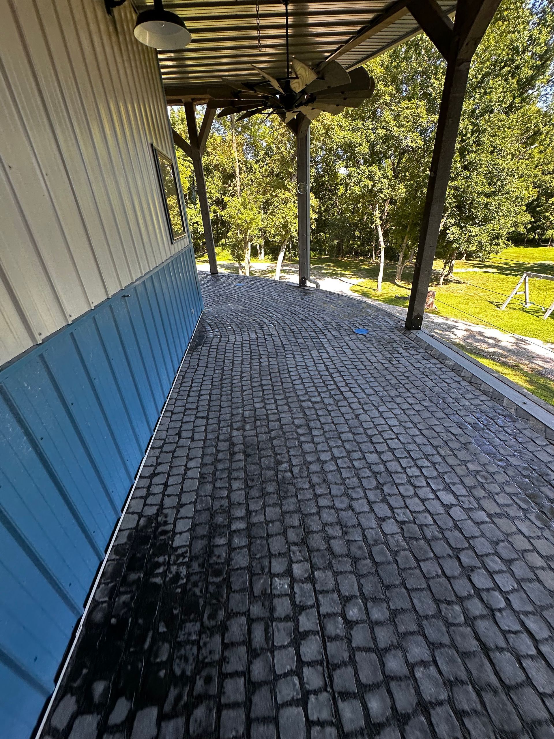 Cobblestone walkway under a shaded overhang next to a blue and white building, with trees in the background.