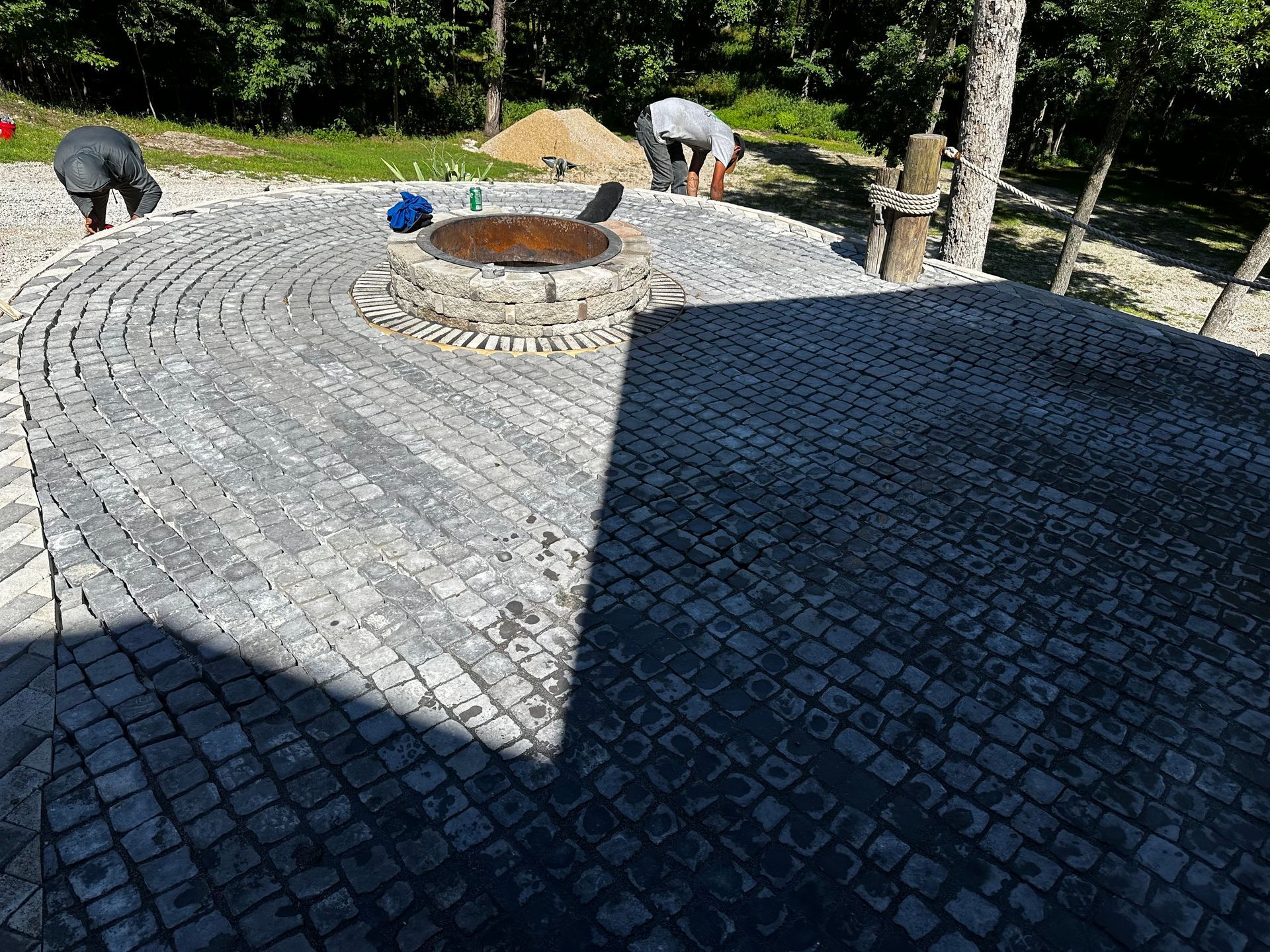 Workers laying cobblestone pavers in a circular pattern around a fire pit outdoors.