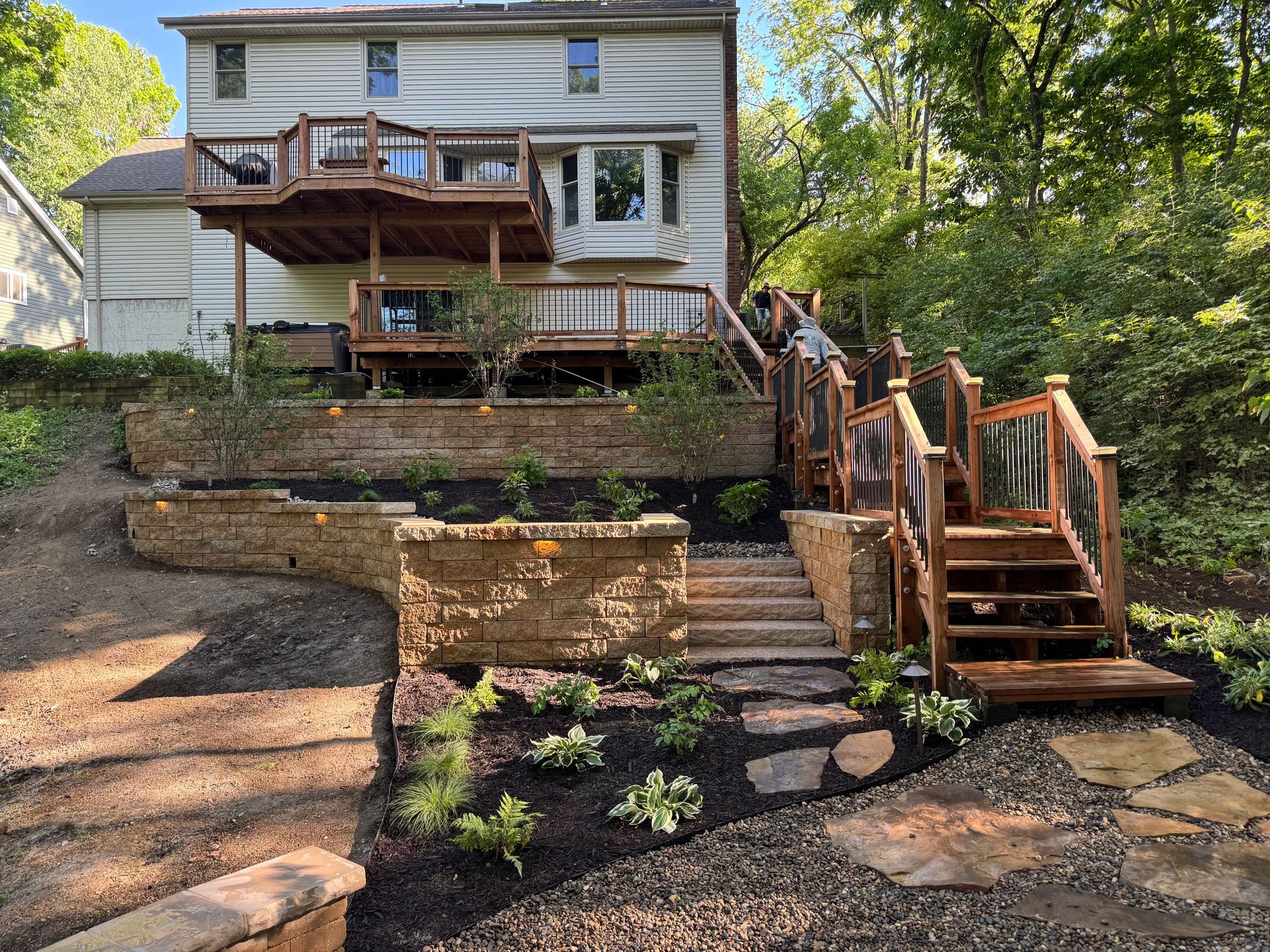 Wooden deck and stairs with glass railings leading up a hill to a two-story house with landscaping.
