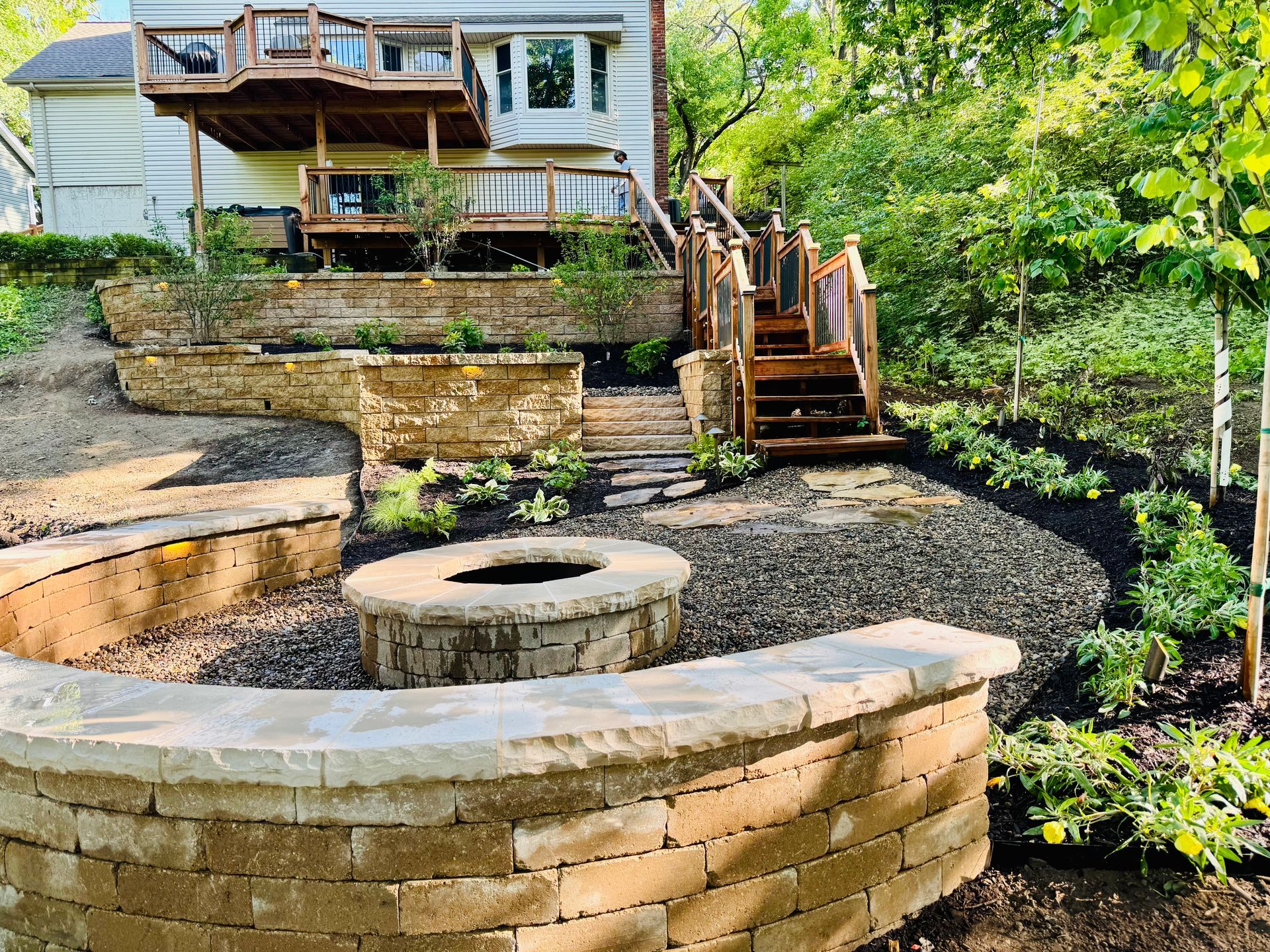 Stone-terraced backyard with built-in seating, fire pit, and stairs leading to a house with a wooden deck.