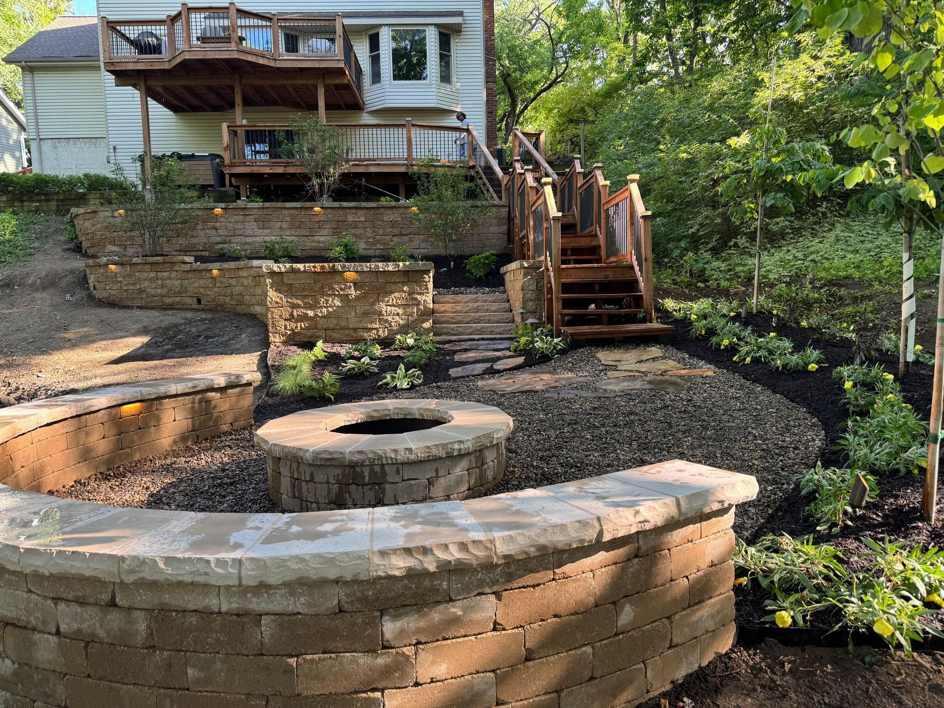 Stone retaining walls and steps leading up to a house with a deck, fire pit in foreground.
