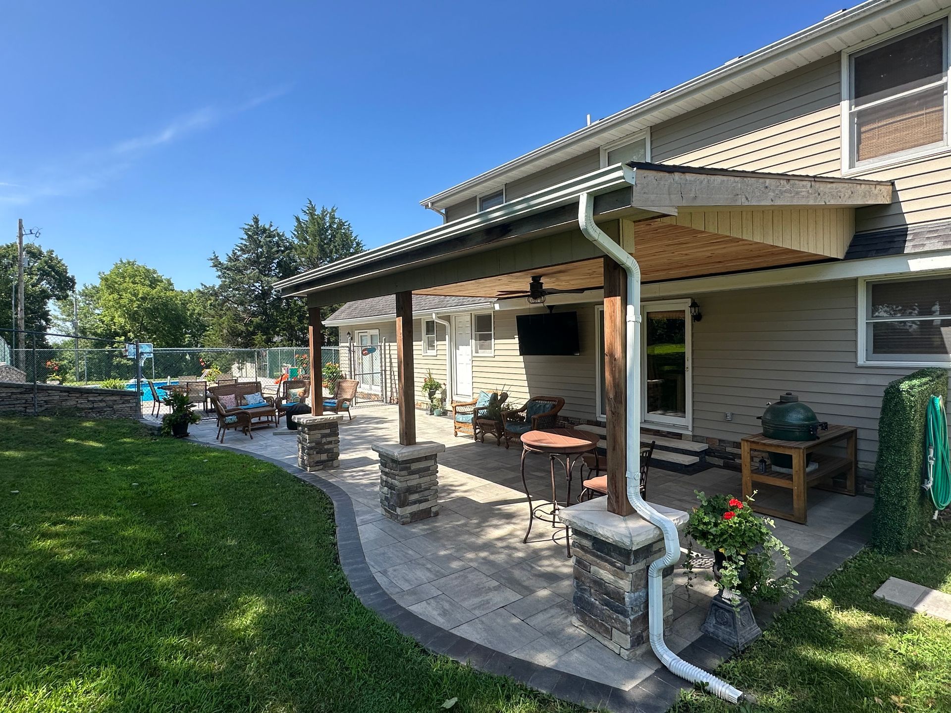 Patio with pavers, covered seating, outdoor TV, and green grass on a sunny day.
