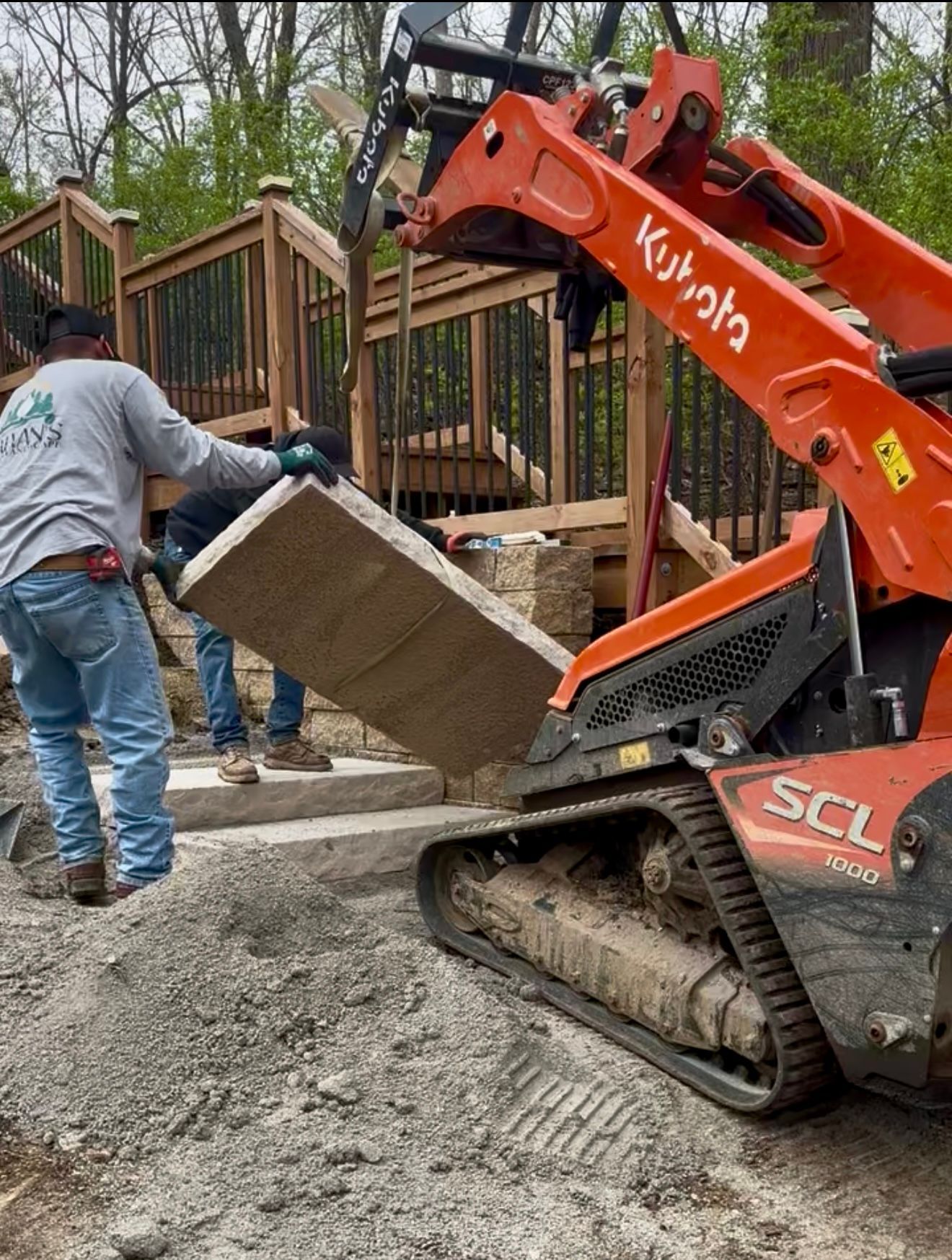 Worker placing a stone slab with a Kubota skid steer near a wooden deck.